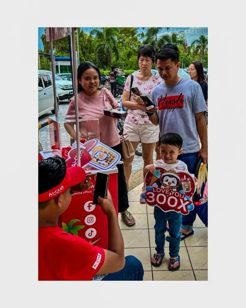 Celebrating Ai Cha’s “Love You 300x” Campaign – Brewing Across Sabah! 8 Ai-CHA staff photographs a smiling child with event sign as parents and visitors gather around the booth