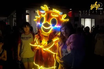 A glowing LED dragon mascot posing with a woman and child during a night celebration at Gamuda Land