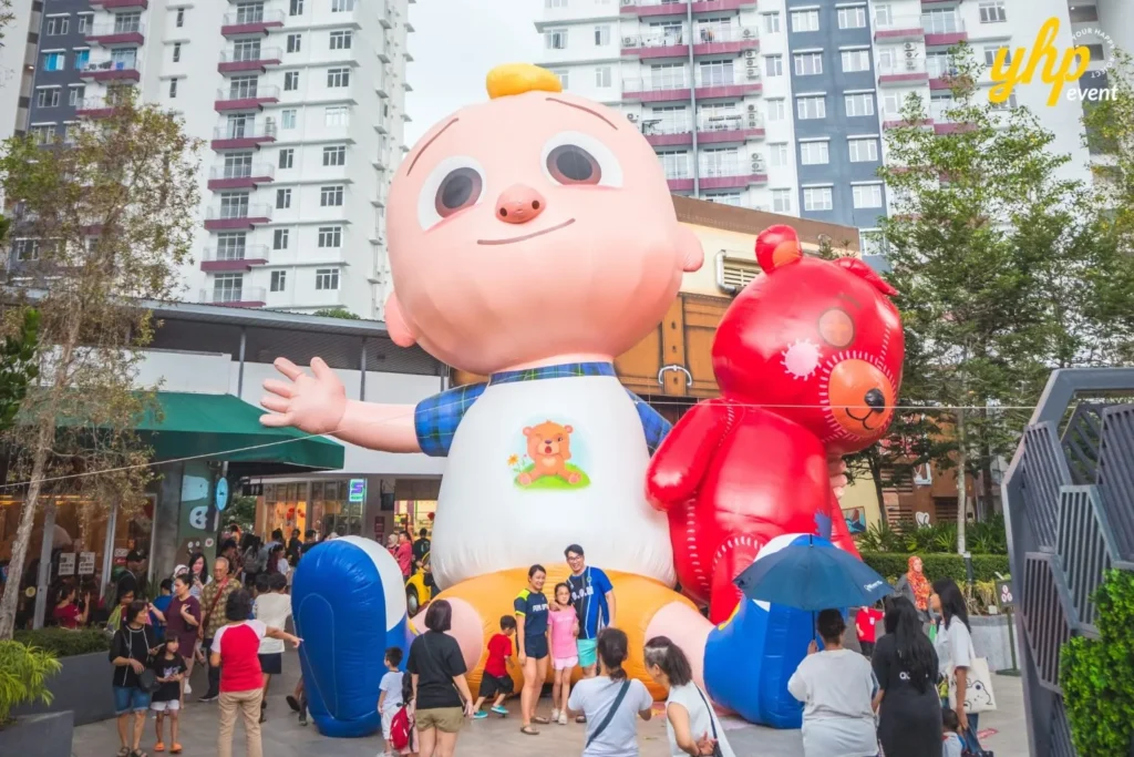 A giant inflatable JJ from CoComelon standing next to a red teddy bear at a busy family carnival