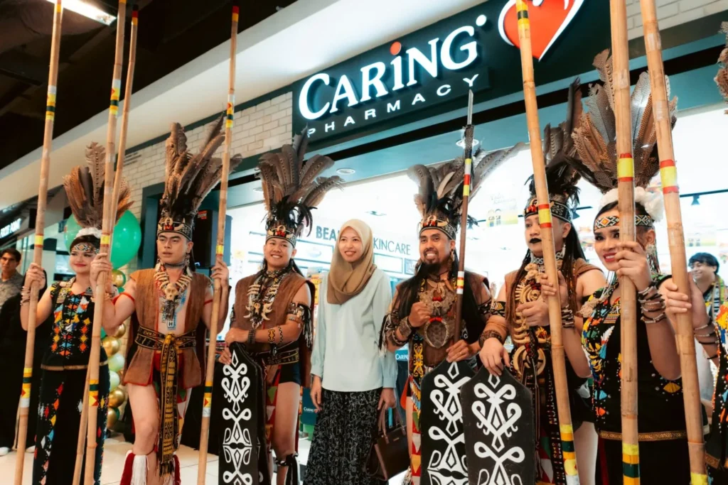 Embrace the Essence of Caring Pharmacy #Promosi Tadau Kaamatan 11 A woman in a hijab posing with Kadazan cultural performers holding tall bamboo poles at Caring Pharmacy