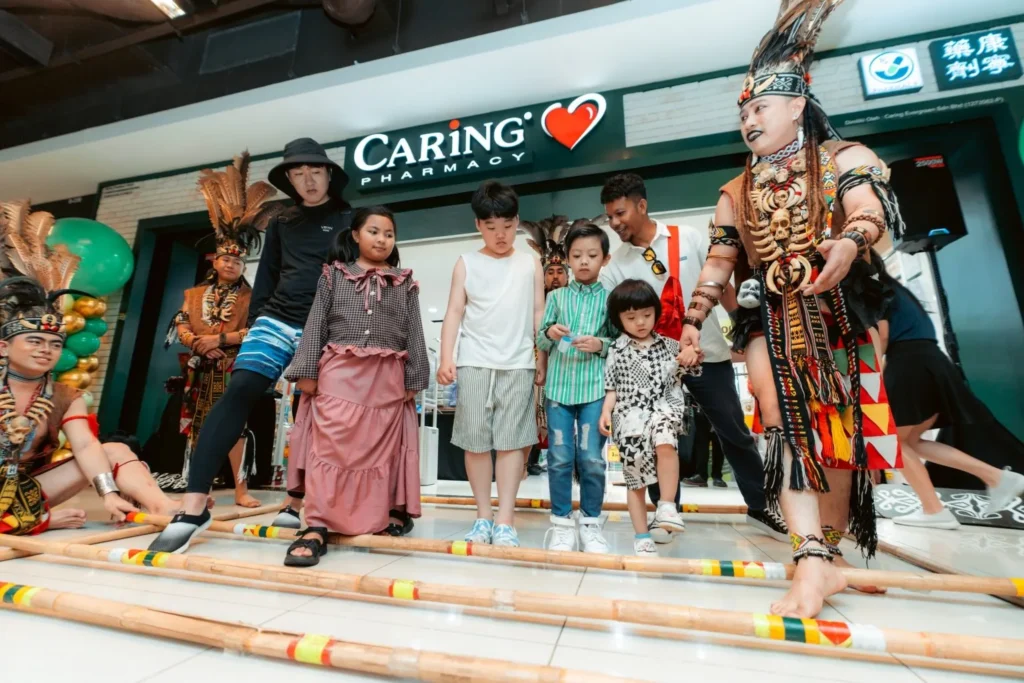 Embrace the Essence of Caring Pharmacy #Promosi Tadau Kaamatan 13 Young children participating in a traditional bamboo Magunatip dance during a Caring Pharmacy event