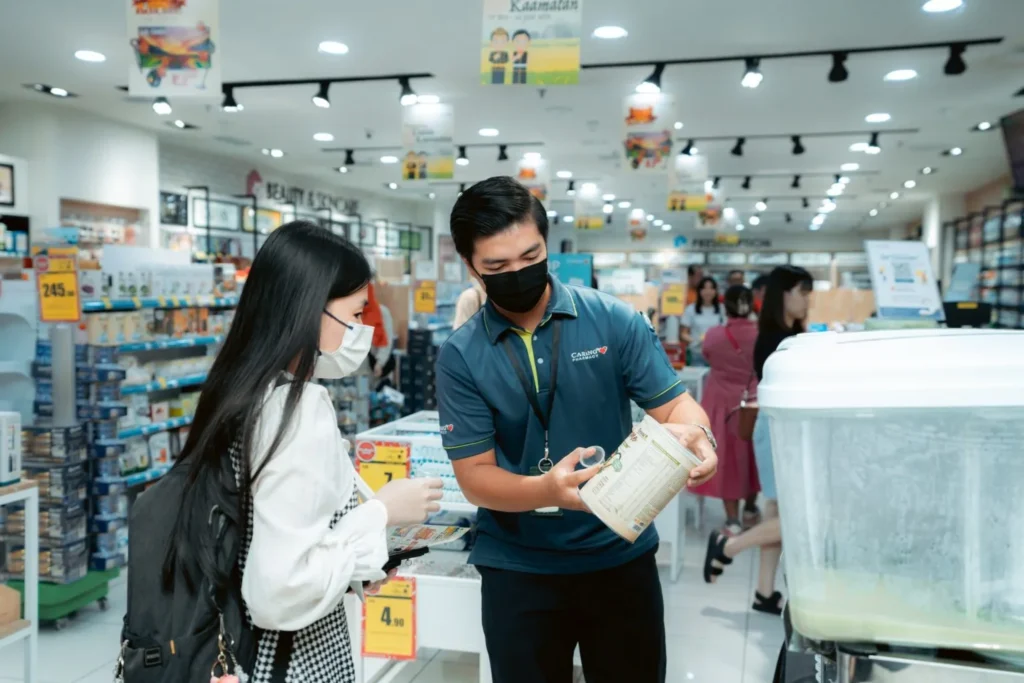 Embrace the Essence of Caring Pharmacy #Promosi Tadau Kaamatan 3 Masked pharmacist in a teal polo assisting a customer with product details at a Caring Pharmacy retail aisle