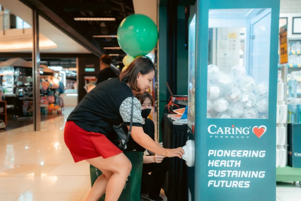 Embrace the Essence of Caring Pharmacy #Promosi Tadau Kaamatan 6 A woman participating in a gashapon capsule prize game at a Caring Pharmacy promotional booth