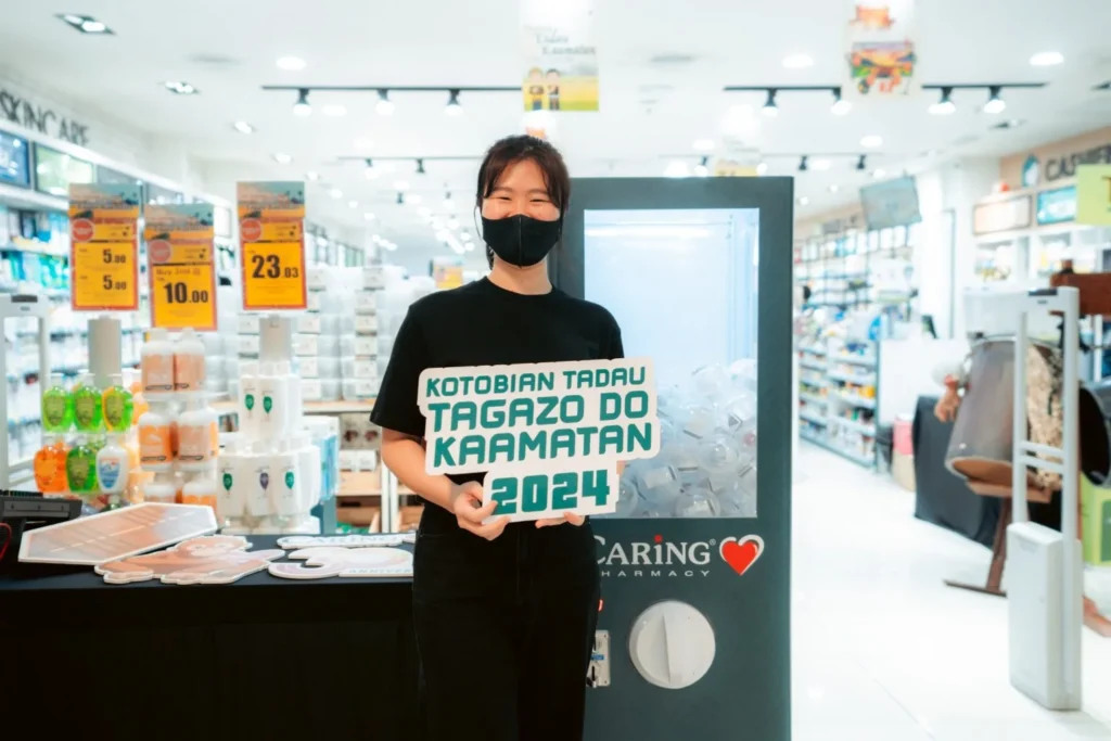 Embrace the Essence of Caring Pharmacy #Promosi Tadau Kaamatan 9 Staff member holding a Kotobian Tadau Tagazo Do Kaamatan 2024 sign at a Caring Pharmacy festive display booth