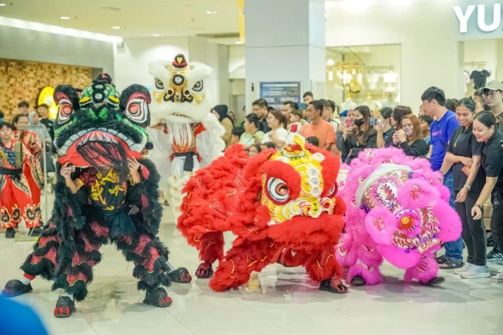Caring Pharmacy Kaamatan & Gawai Campaign Launch 7 Crowd gathers around red, pink, white and black lion dance performers at a mall event outside the pharmacy