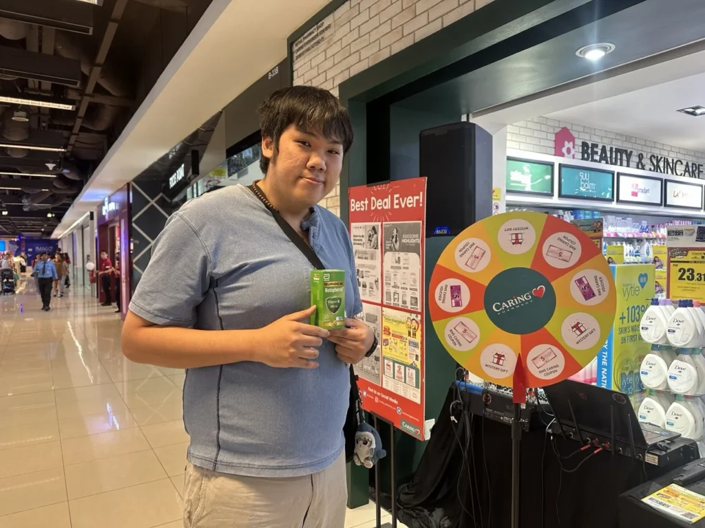 Caring Pharmacy Kaamatan & Gawai Campaign Launch 9 Shopper holding a product beside a Caring Pharmacy prize wheel during an in-store event display at Imago mall