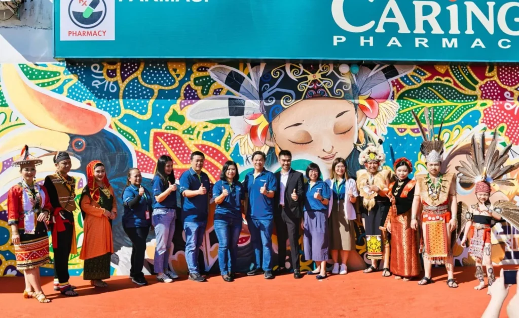 Caring Pharmacy Mural Unveiling l Kota Sentosa Kuching 2 Group photo in front of Caring Pharmacy Kota Sentosa Kuching mural with staff and cultural performers in traditional attire outdoors