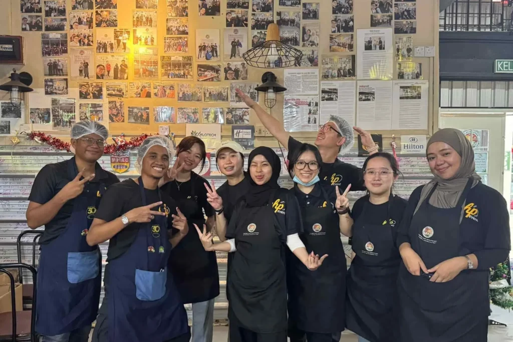 Dapur Kita-Kita: Sepinggan Kasih, Sebakul Harapan 4 Volunteer team in aprons and hairnets posing together inside community kitchen beneath a wall of event photos