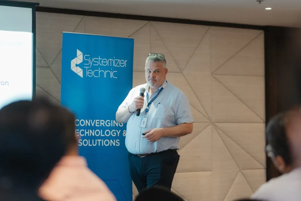 Ignite IT : Powering Tomorrow’s Solutions 4 A male speaker presenting technical insights at a Systemizer Technic corporate event with blue banners