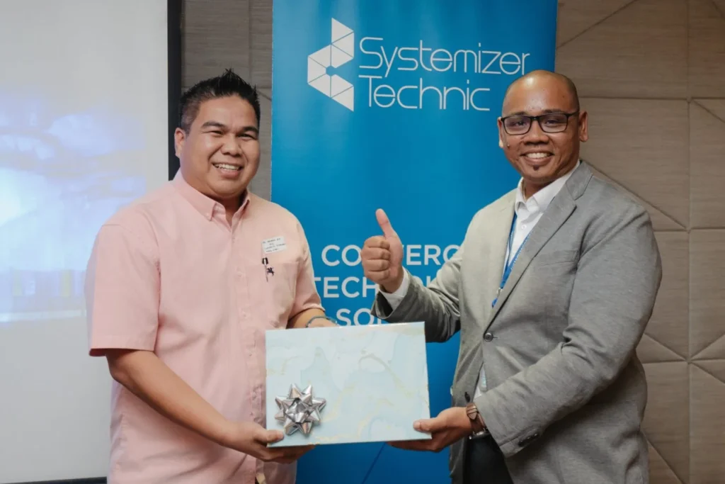 Ignite IT : Powering Tomorrow’s Solutions 6 Two men smiling and holding a gift box in front of a blue Systemizer Technic brand banner at an event
