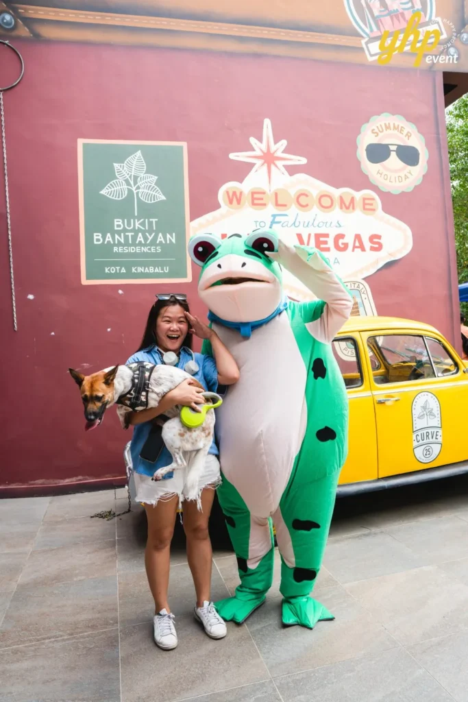 A woman holding a cattle dog standing next to a person in a green frog mascot costume