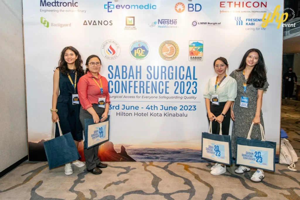 Exploring Advancements in Surgical Care: Highlights from the Sabah Surgical Conference 3 Attendees holding event tote bags in front of the Sabah Surgical Conference 2023 backdrop at Hilton Kota Kinabalu