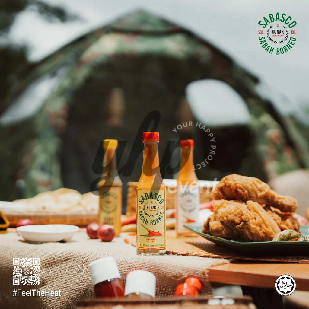 The Local Sabah Chili Sauce Brand That’s Heating Up 11 Sabasco Sabah Borneo chili sauce bottles displayed on a picnic table with fried chicken and a tent in the background