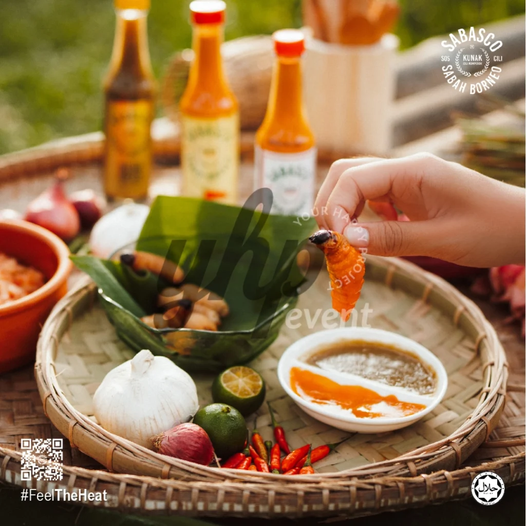 The Local Sabah Chili Sauce Brand That’s Heating Up 14 Hand holding a cooked sago grub above dipping sauce, herbs, and Sabasco Sabah Borneo chili sauce on a woven tray