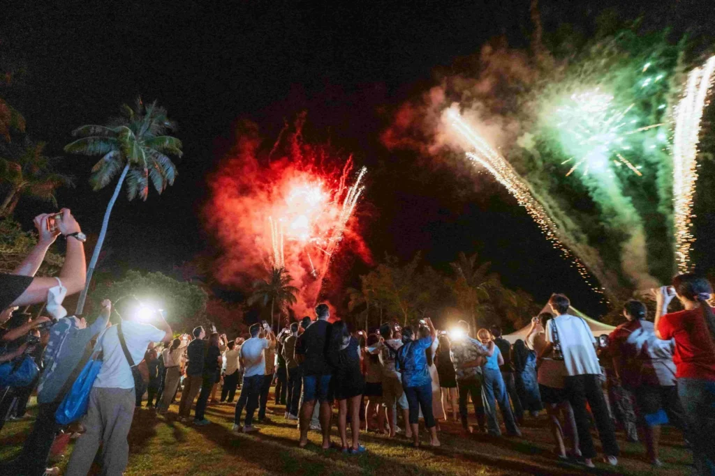 Spark Fest Carnival l Karambunai Hotel 8 Crowd watching red and green fireworks light up the night sky over palm trees during an outdoor beachside event