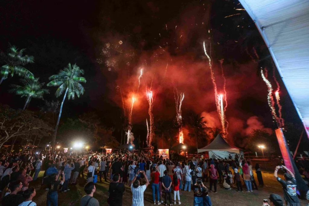 Spark Fest Carnival l Karambunai Hotel 2 Large crowd photographs red fireworks rising above palm trees and tents at an open air evening festival at Karambunai Hotel.