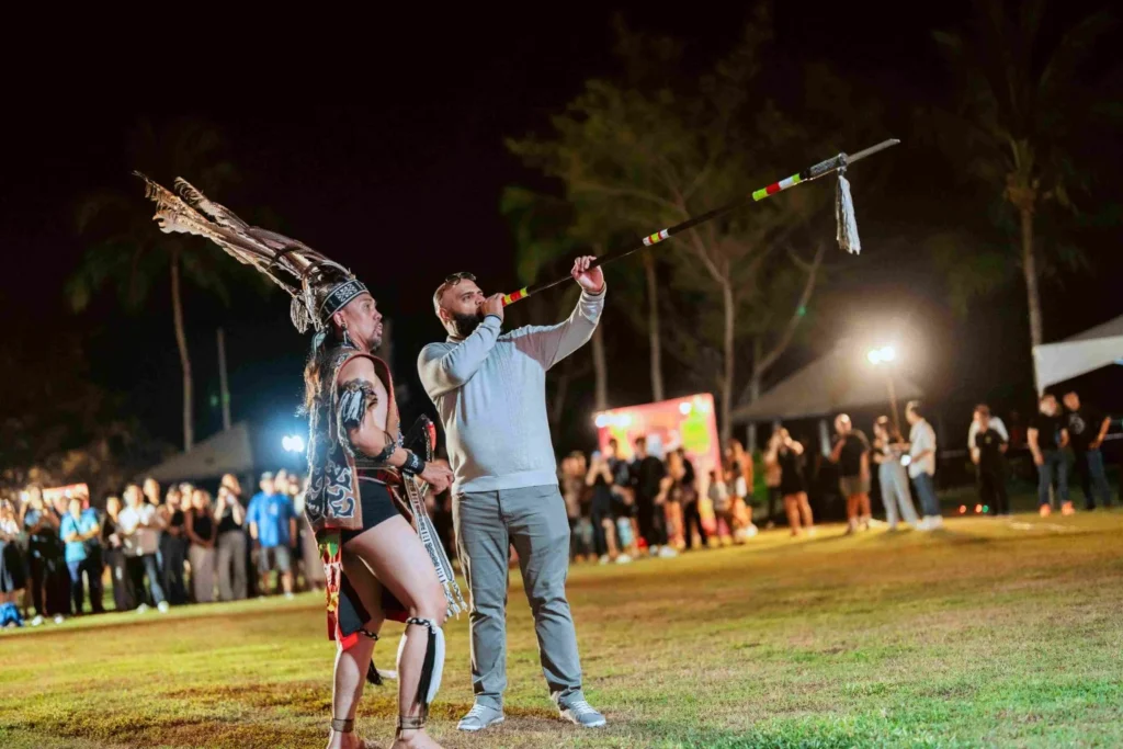 Spark Fest Carnival l Karambunai Hotel 3 Man blows a ceremonial horn beside a Sabah traditional cultural performer on a field during a nighttime festival
