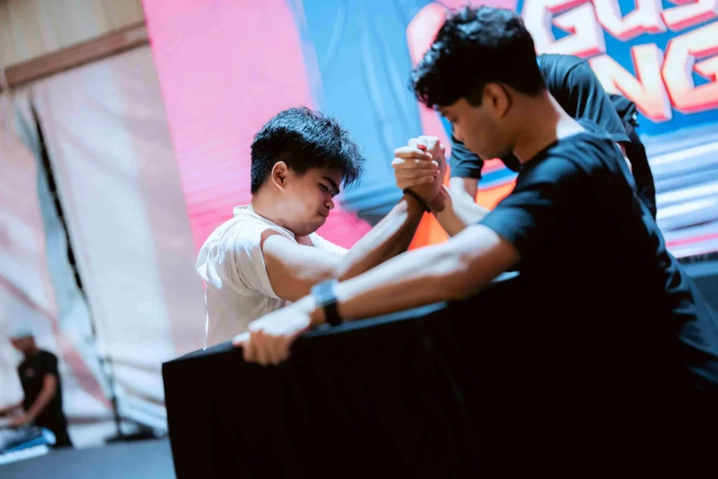 Spark Fest Carnival l Karambunai Hotel 4 Two men compete in an arm wrestling match on stage under colorful lights during an indoor carnival event at Karambunai Hotel