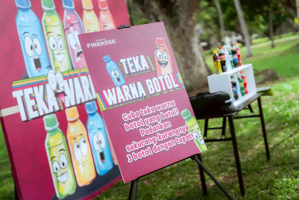 Spark Fest Carnival l Karambunai Hotel 7 Signboard for a bottle color guessing game stands beside arranged bottles on tables in a shaded park area