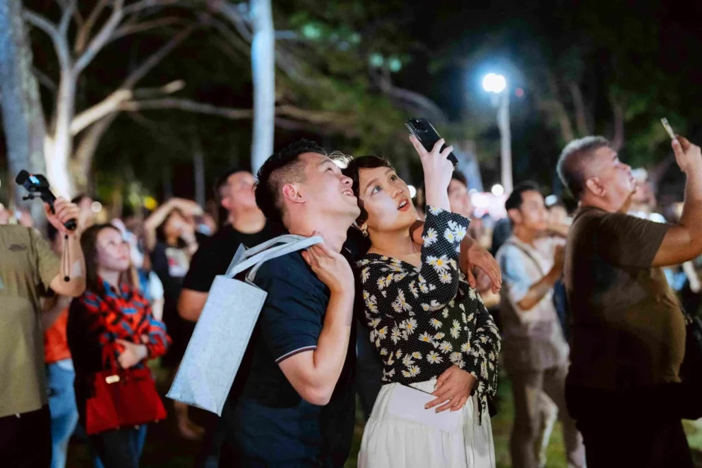 Spark Fest Carnival l Karambunai Hotel 9 Couple looks up and films the night sky as a crowd gathers to watch an outdoor fireworks show nearby