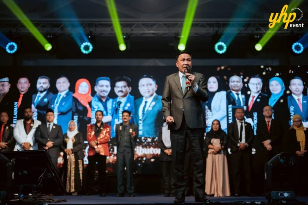 TIENS Sabah Convention l Borneo Royale Hotel Tawau 3 TIENS host speaks at center stage as award recipients and event officials stand before a lit video backdrop in Tawau , Sabah