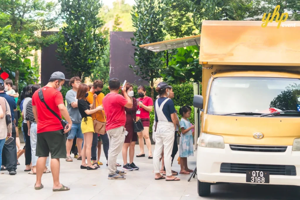 #TooHuattoHandle Lunar New Year Celebration 16 Visitors queue beside a yellow food truck at an outdoor community market surrounded by trees and family-friendly stalls